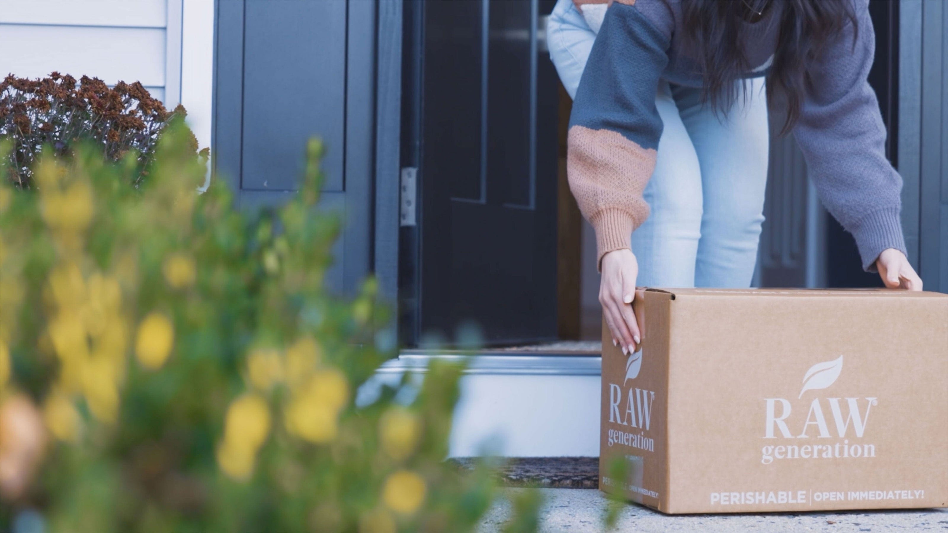 A woman stands outdoors holding a box, wearing jeans, with a building partially visible in the background.