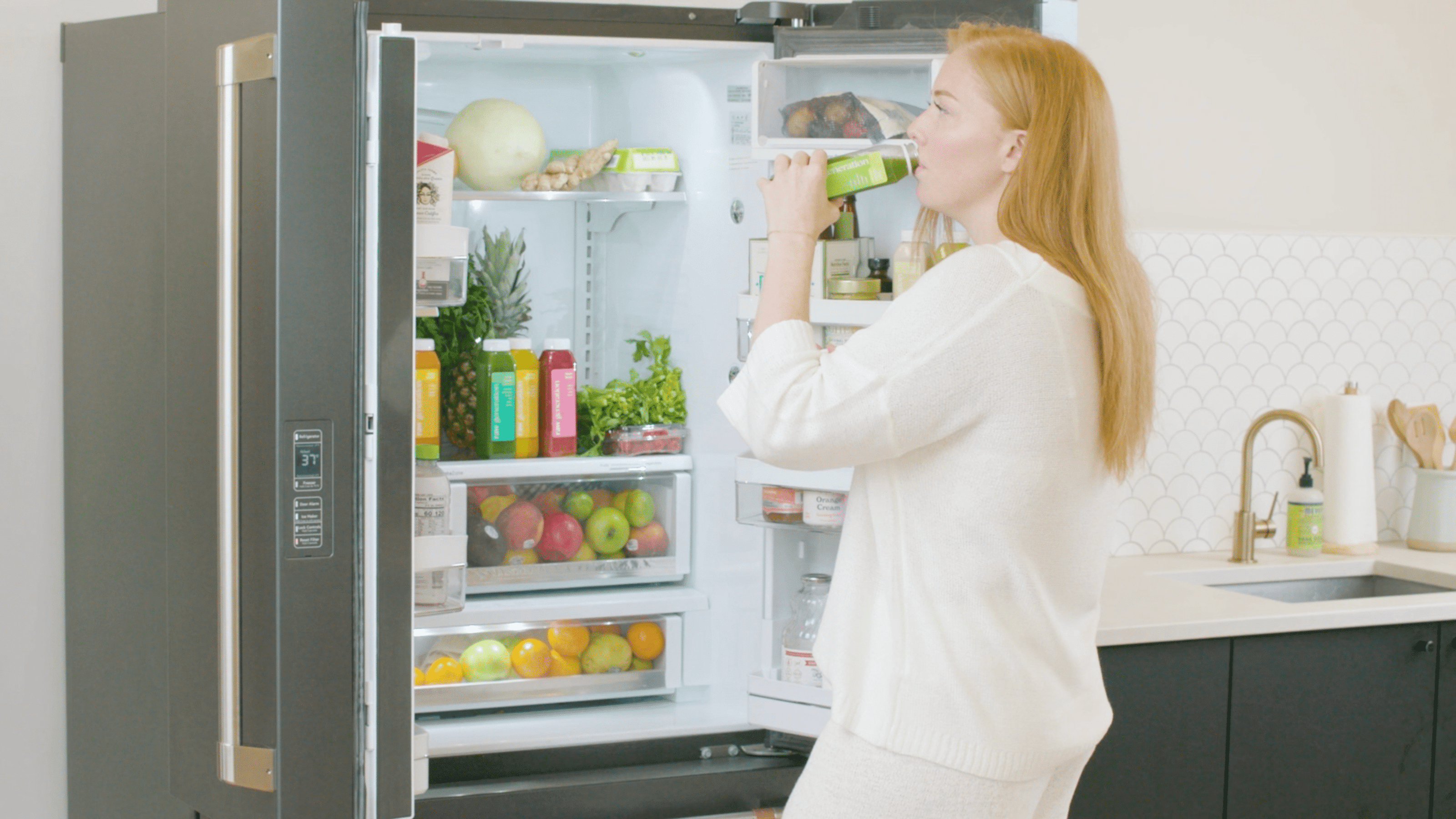 A woman drinks from a green bottle in front of an open refrigerator, with visible fruits and a pink box inside.