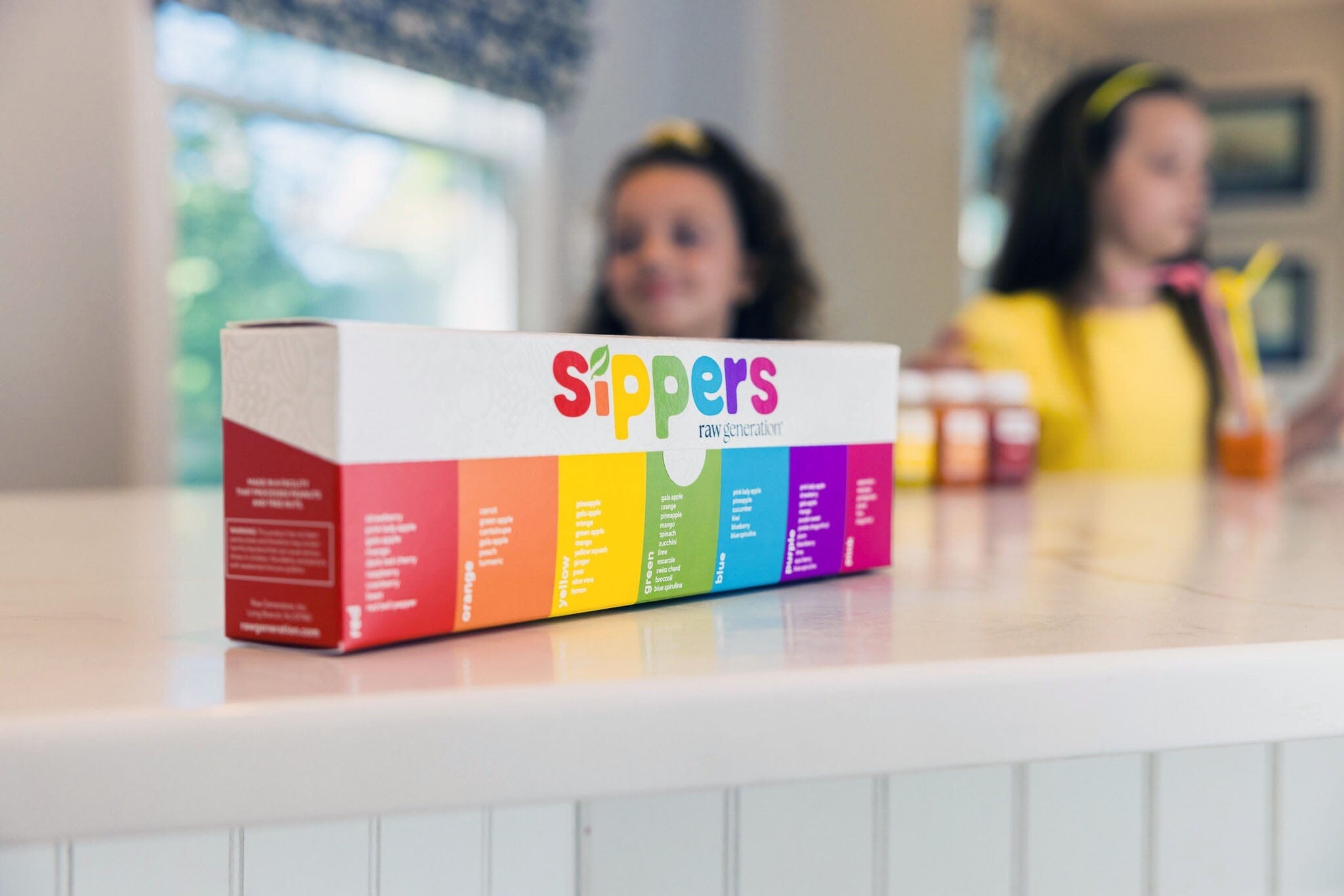 Colorful box of sippers on a counter, partially obscured by blurred images of a girl with a juice bottle and a woman's face in the background.