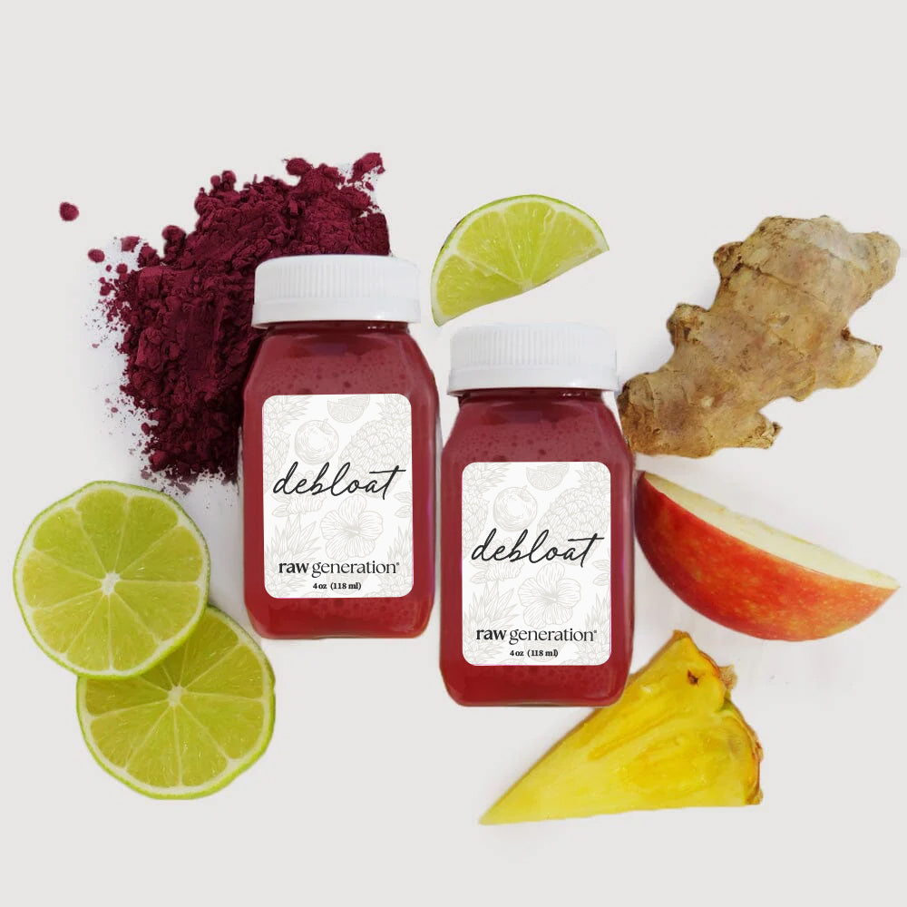 Two 4-oz plastic bottles of Bridal De-Bloat juice shots, surrounded by sliced lime, pineapple, apple, ginger, and a pile of deep-red powder.