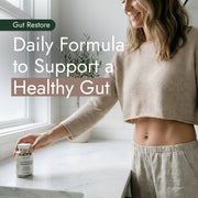 Woman in loungewear reaching for Gut Restore Daily Debloat Formula capsules on a kitchen countertop, promoting gut health support and daily digestive wellness naturally.