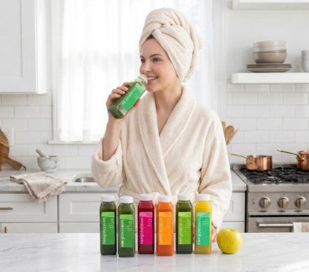 Beauty Box™ juices lined up on a kitchen counter, showcasing vibrant colors. A person sips a green juice, emphasizing the skincare routine's nourishing benefits.