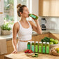 Woman in kitchen with Daily Detox Box, sipping green juice among bottles, strawberries, cucumber, apples, kale, ginger, lemons, emphasizing wellness and natural cleansing.
