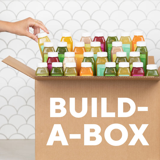 A cardboard box of vibrant, cold-pressed juice bottles is enticingly showcased as a hand selects one from the top row. BUILD-A-BOX by Raw Generation is boldly displayed on the side, set against a white tiled wall that suggests refreshing choices for gut health.